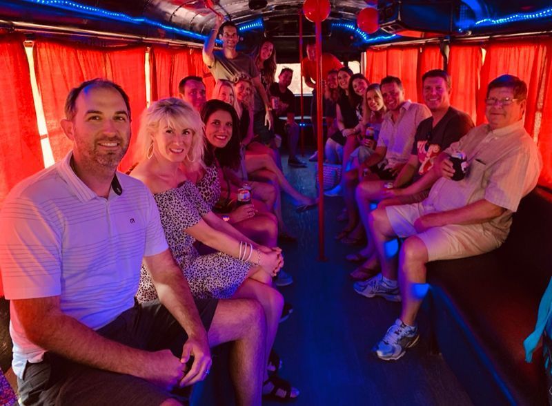 Guests seated along the bench seating of a party bus with red curtains and blue LED lighting.